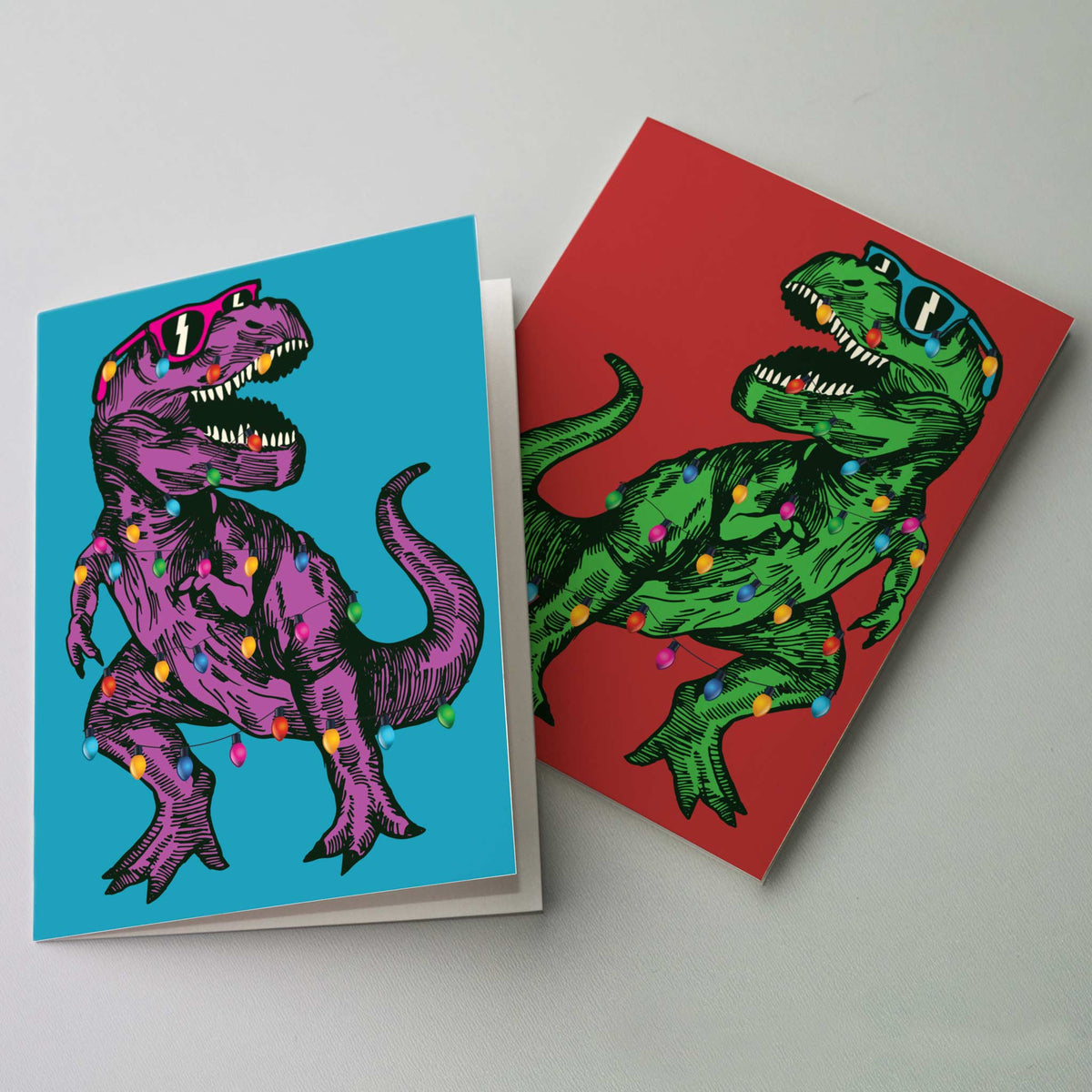 Christmas cards featuring a hatched T-Rex in sunglasses wrapped in multicolor string lights, two colorways.