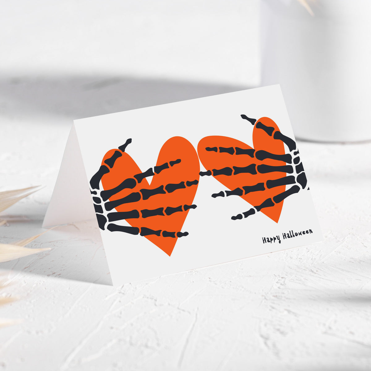 Halloween card set with two skeletal hands, each resting on a solid orange heart, script text below.