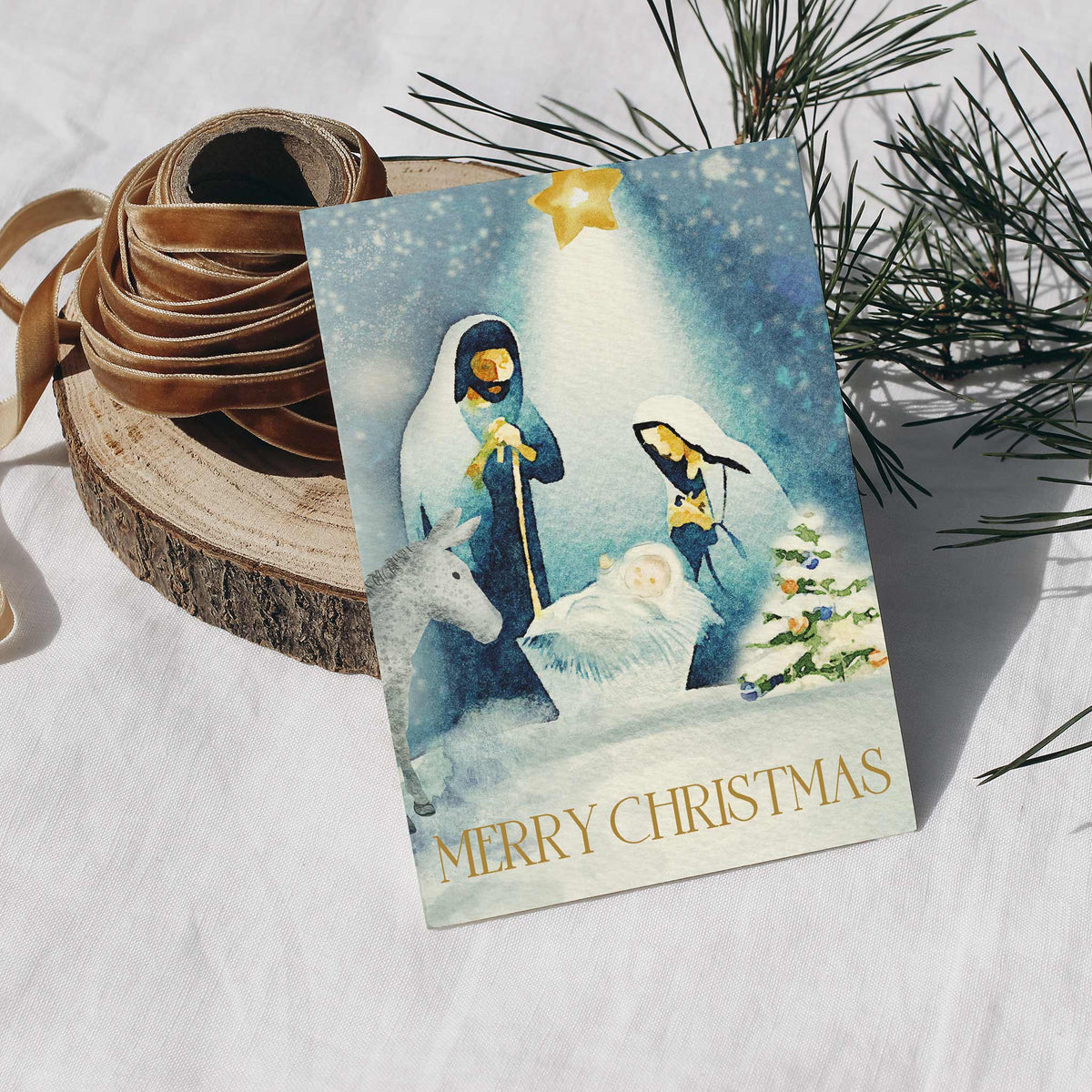 Watercolor nativity scene card with robed figures, infant in white cloth, gray donkey, and snow-covered ground.