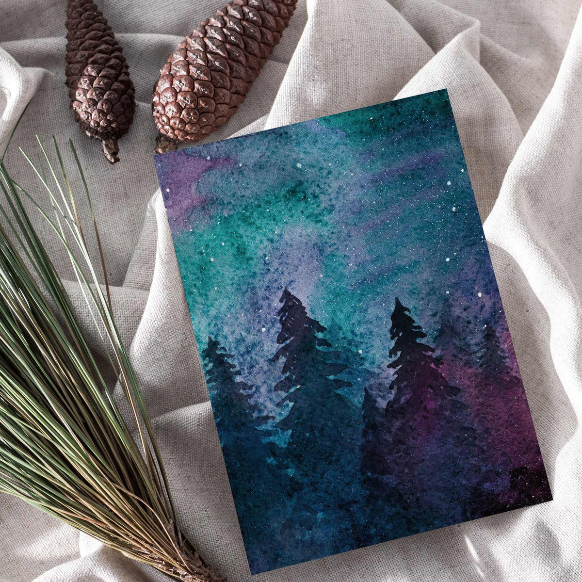 Holiday card front panel showing black evergreen tree silhouettes against a colorful watercolor sky wash.