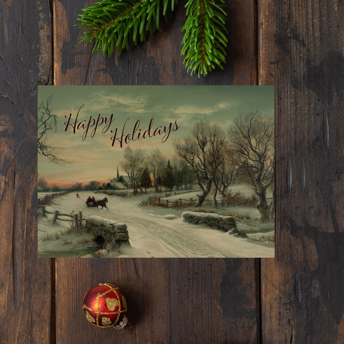 Reproduction chromolithograph holiday card, snowy road, sleigh, bare trees, church spire, "Happy Holidays" in maroon script.