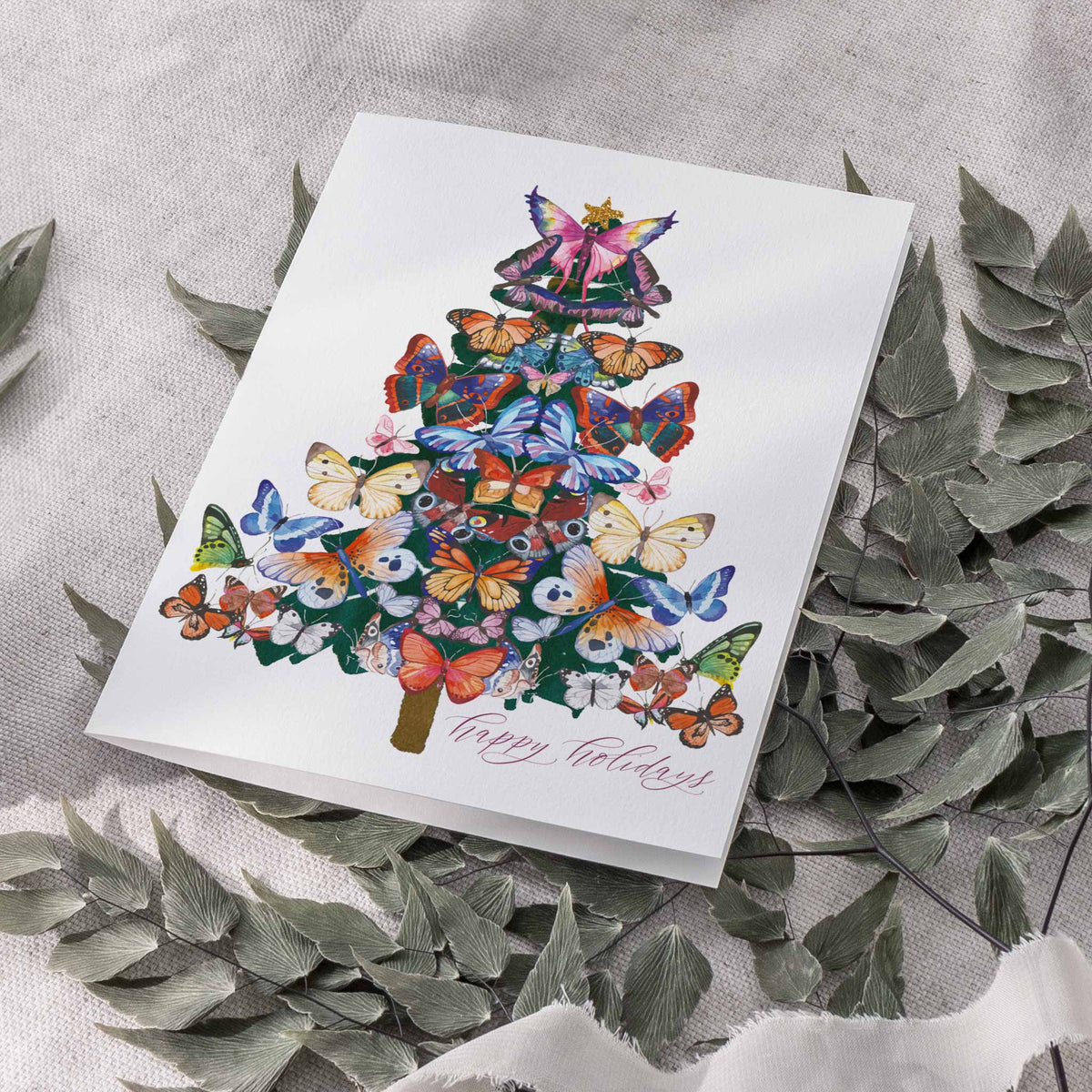 Illustrated card with a butterfly Christmas tree over green foliage and a brown trunk, cursive text at lower right.