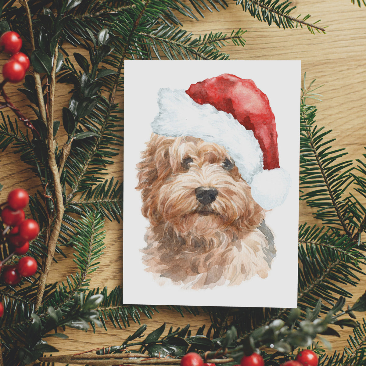 Golden curly-haired labradoodle painted in watercolor, wearing a red Santa hat with white pompom, portrait card.