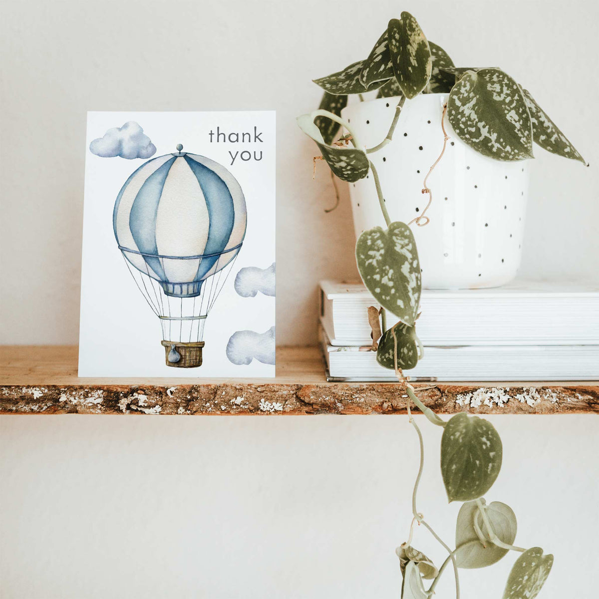 Watercolor balloon with small hanging teardrop detail and three rounded clouds; "thank you" in charcoal gray lowercase text.