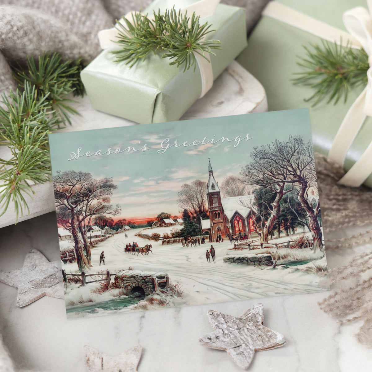 Holiday cards depicting a twilight village scene with a lit church, sleigh passengers, and script greeting.