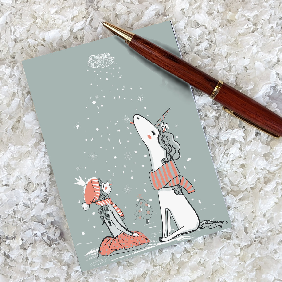 Winter cards, 24 pack, featuring a girl and white unicorn in matching striped scarves beside a small tree in falling snow.